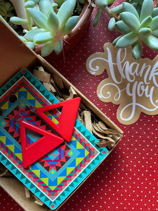 Red authentic 80s geometric earrings in a box with colorful packaging, next to a 'thank you' card on a red retro patterned surface.