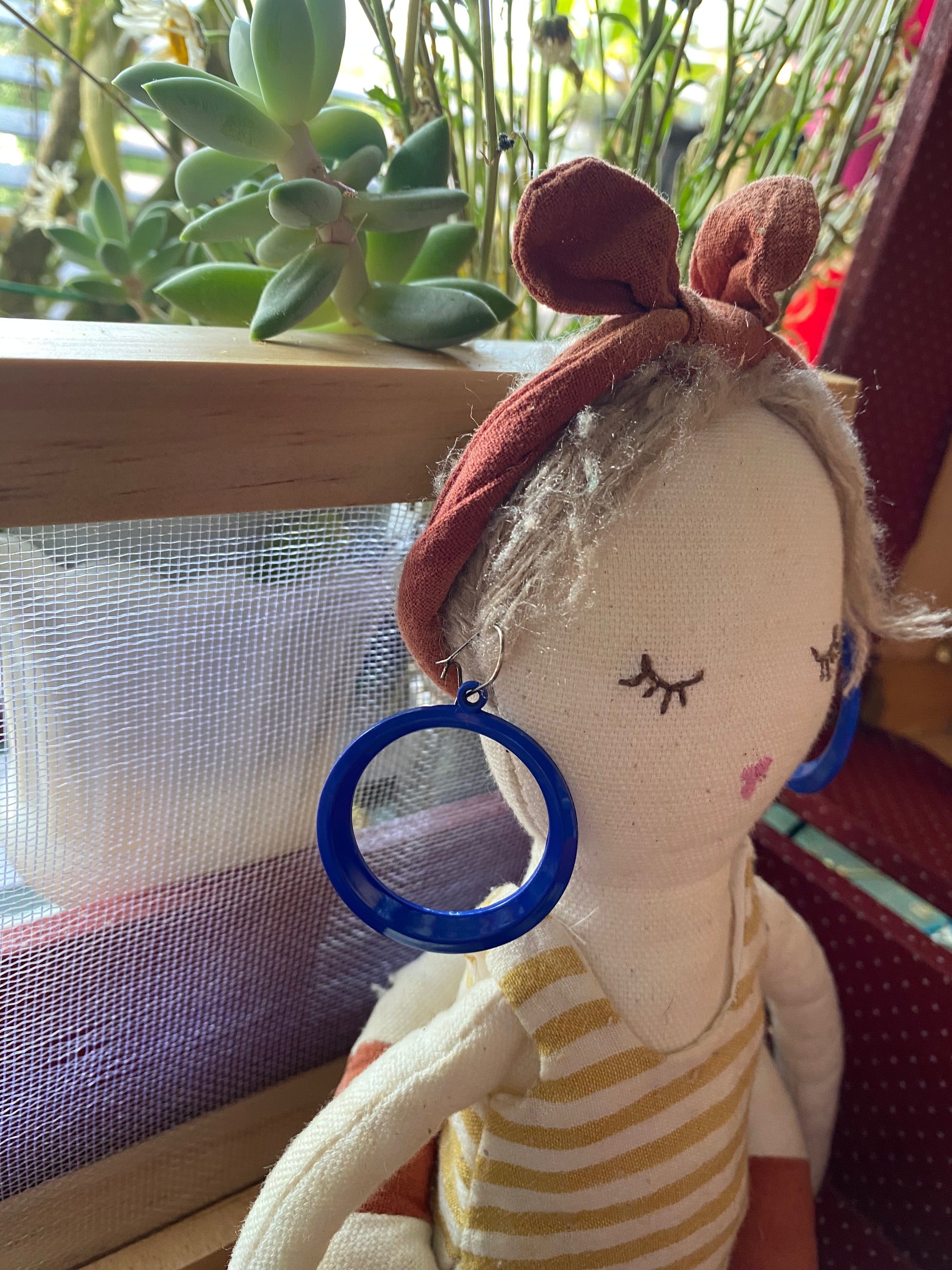 Fabric doll with a blue 80s hoop earring in front of a window with plants
