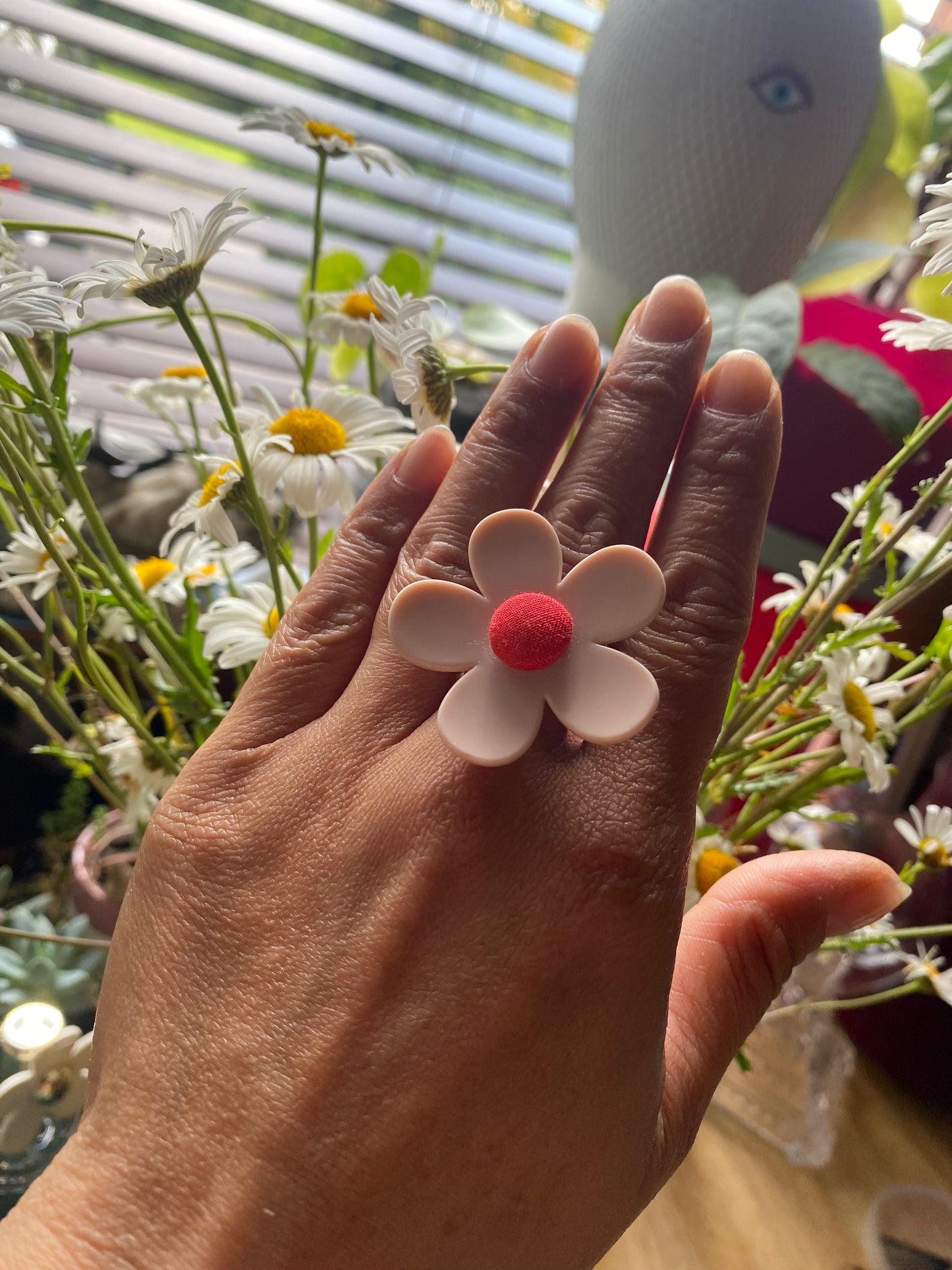 Hand wearing a handmade daisy flower-shaped ring with a red center, surrounded by flowers.