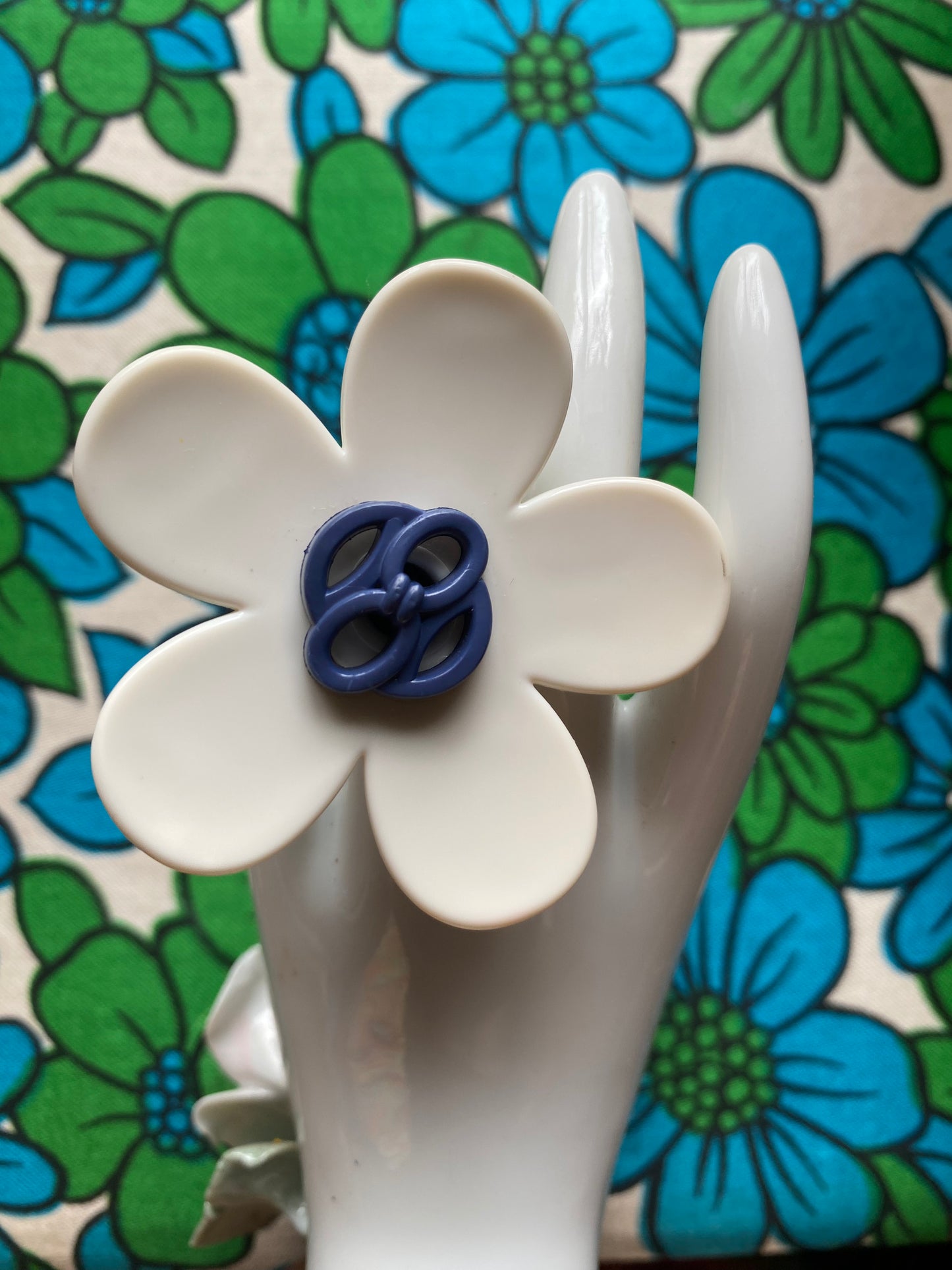 White ceramic hand wearing a handmade daisy flower with blue center against a retro floral background