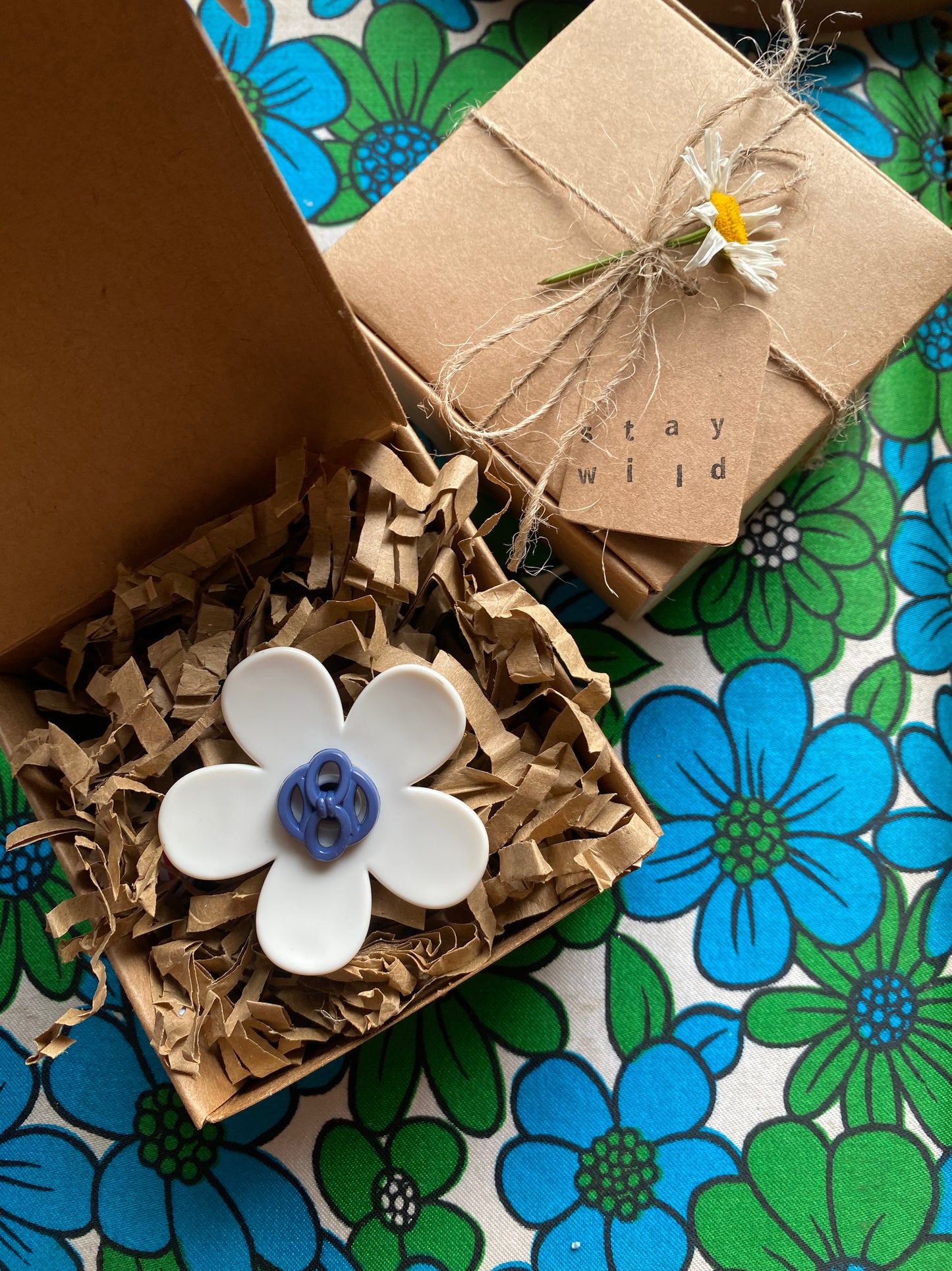 White handmade daisy flower ring with blue center in a eco friendly brown paper box on a retro floral-patterned surface