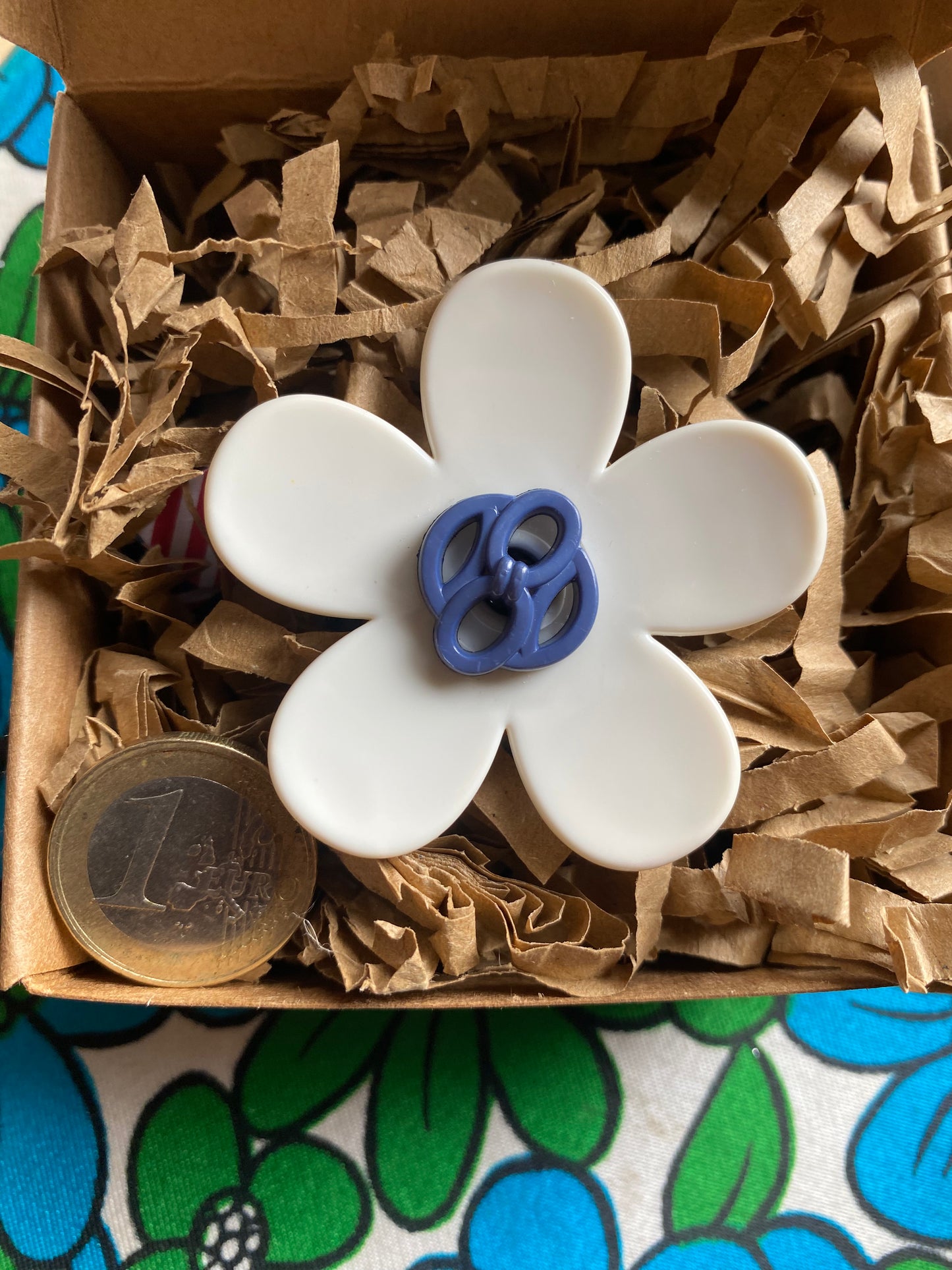 White handmade daisy flower-shaped ring with a blue center in a box of brown paper