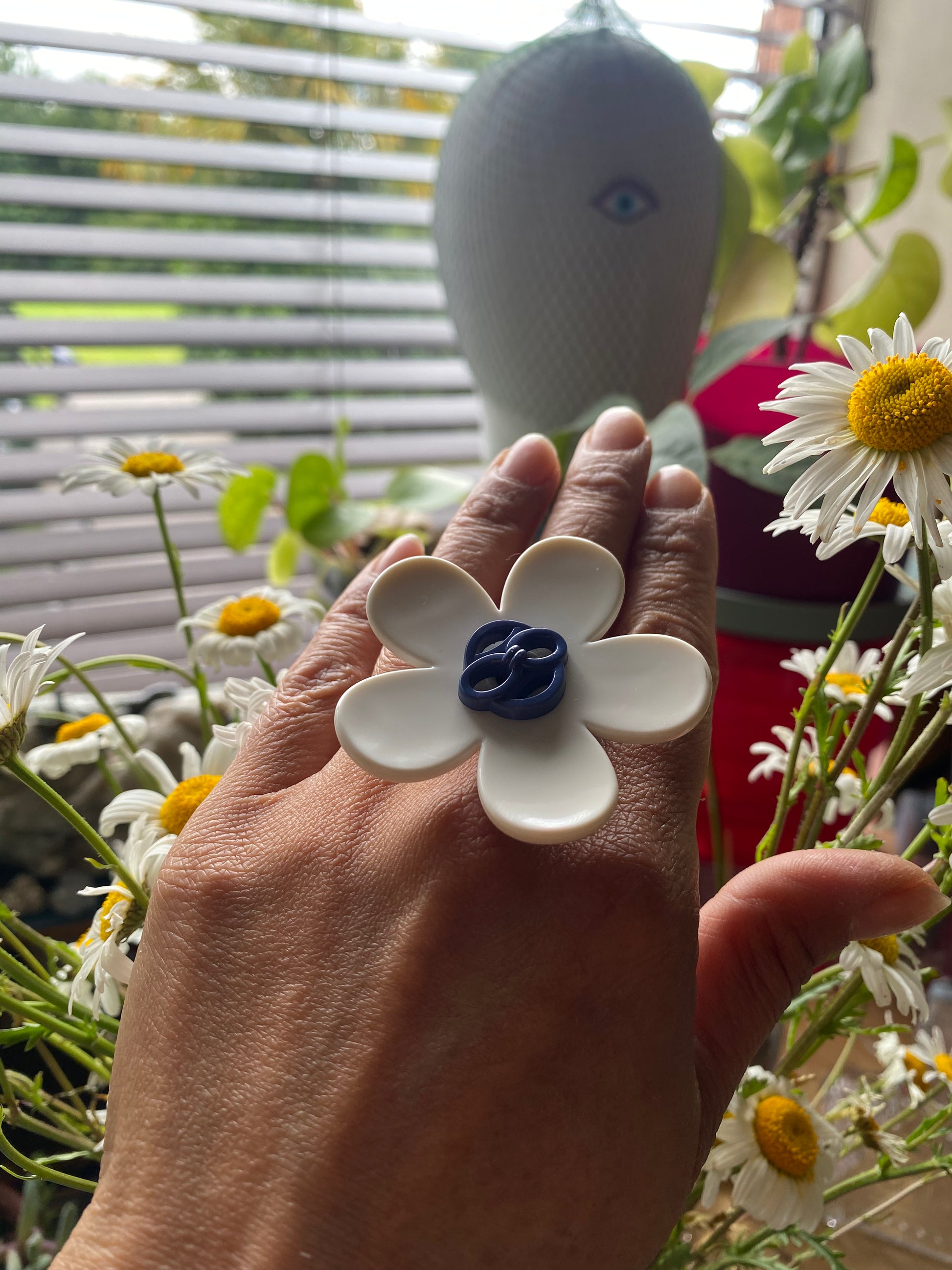 Hand wearing a white handmade daisy flower-shaped ring with a blue center, surrounded by flowers and a blurred background.