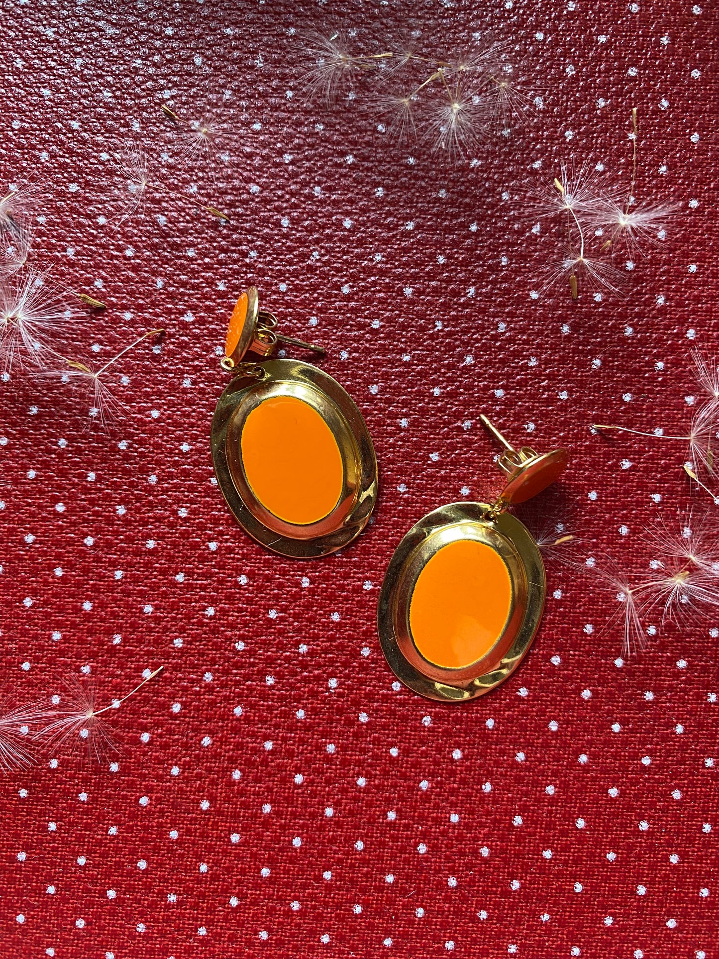 Orange 80s earrings with gold rims on a red fabric background with white polka dots and dandelion flowers.