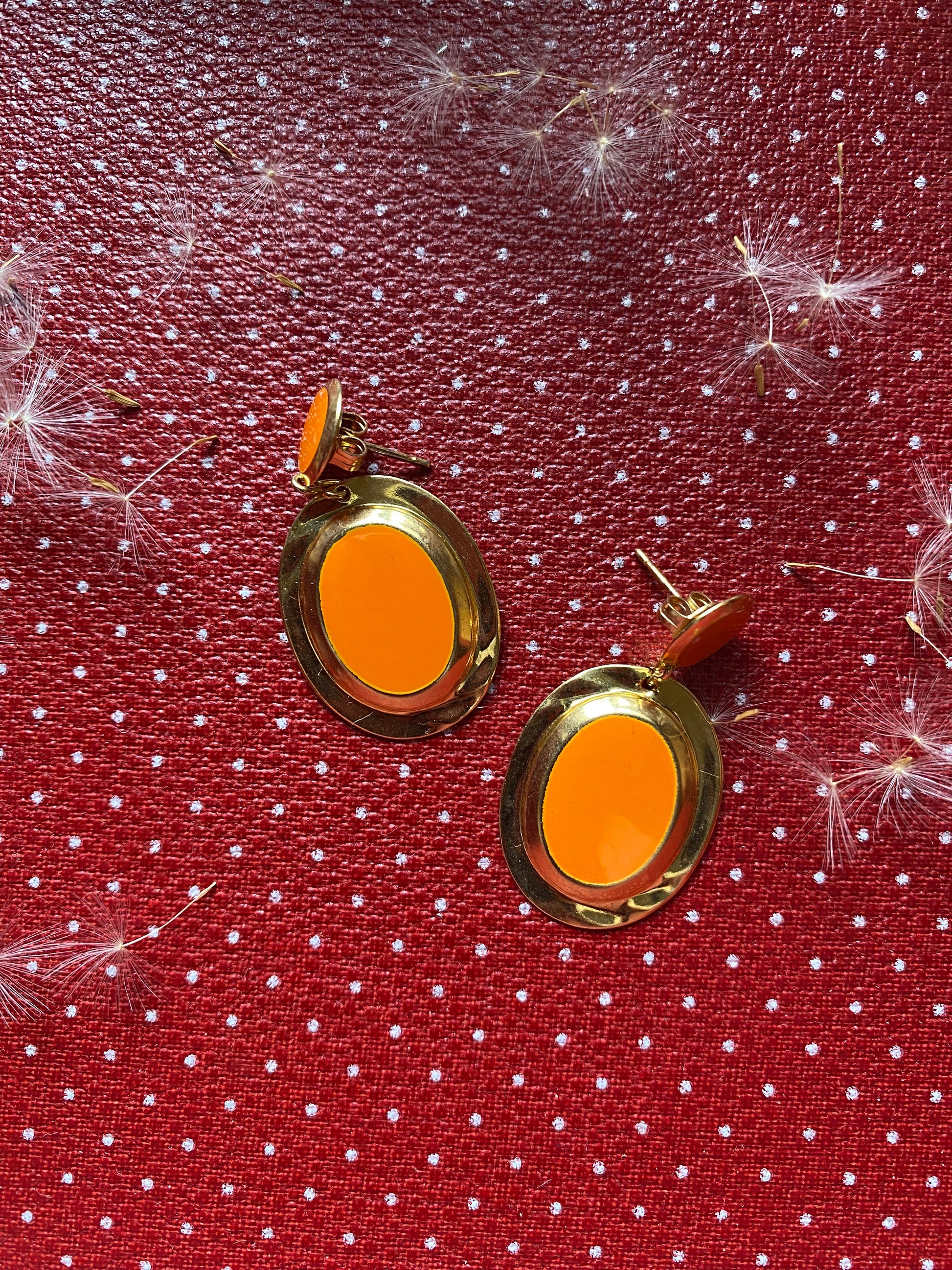 Orange 80s earrings with gold rims on a red fabric background with white polka dots and dandelion flowers.