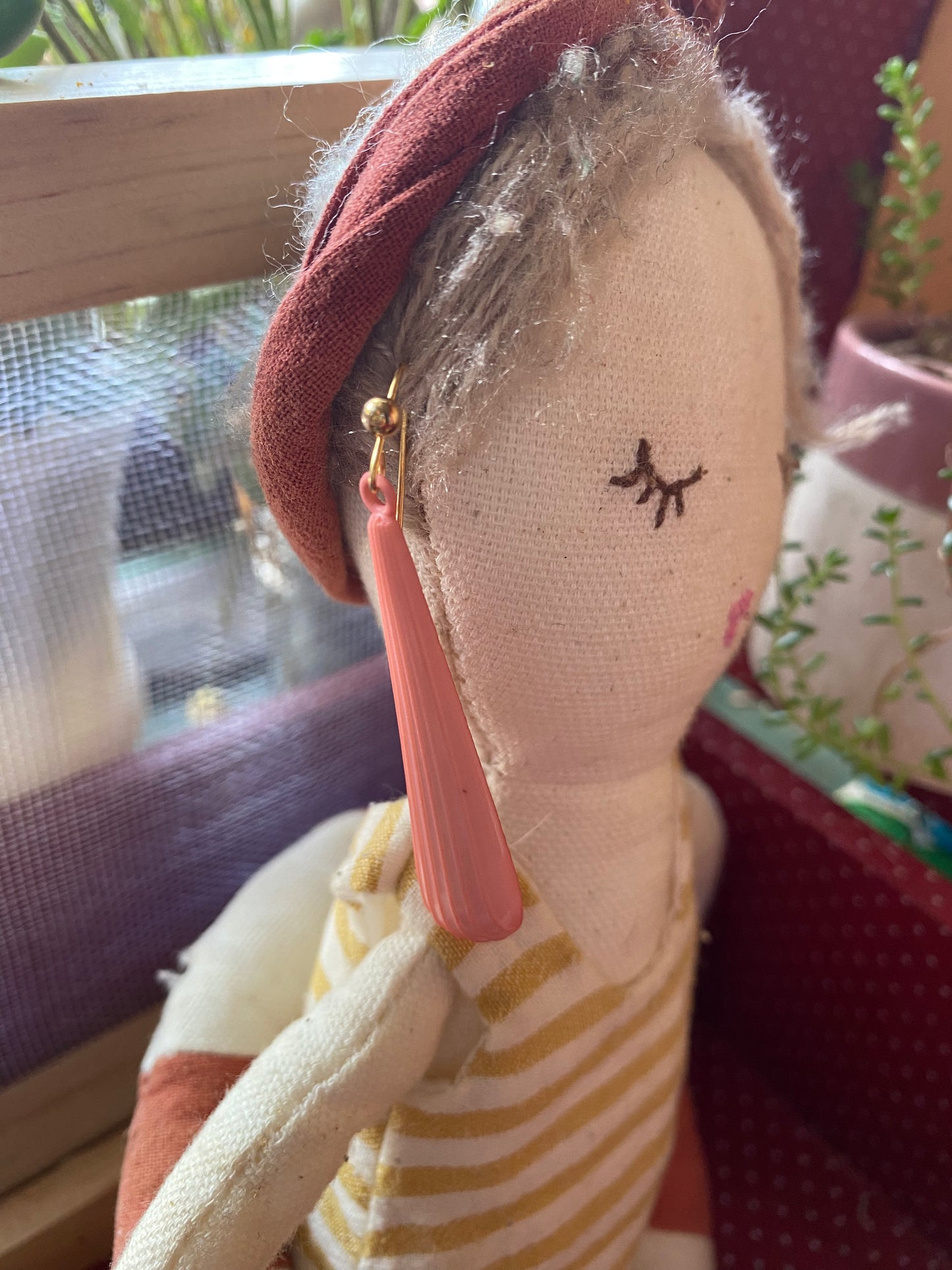 Textile doll modelling the earrings with a red headband and striped dress, held by a pink ribbon, against a blurred indoor background. 
