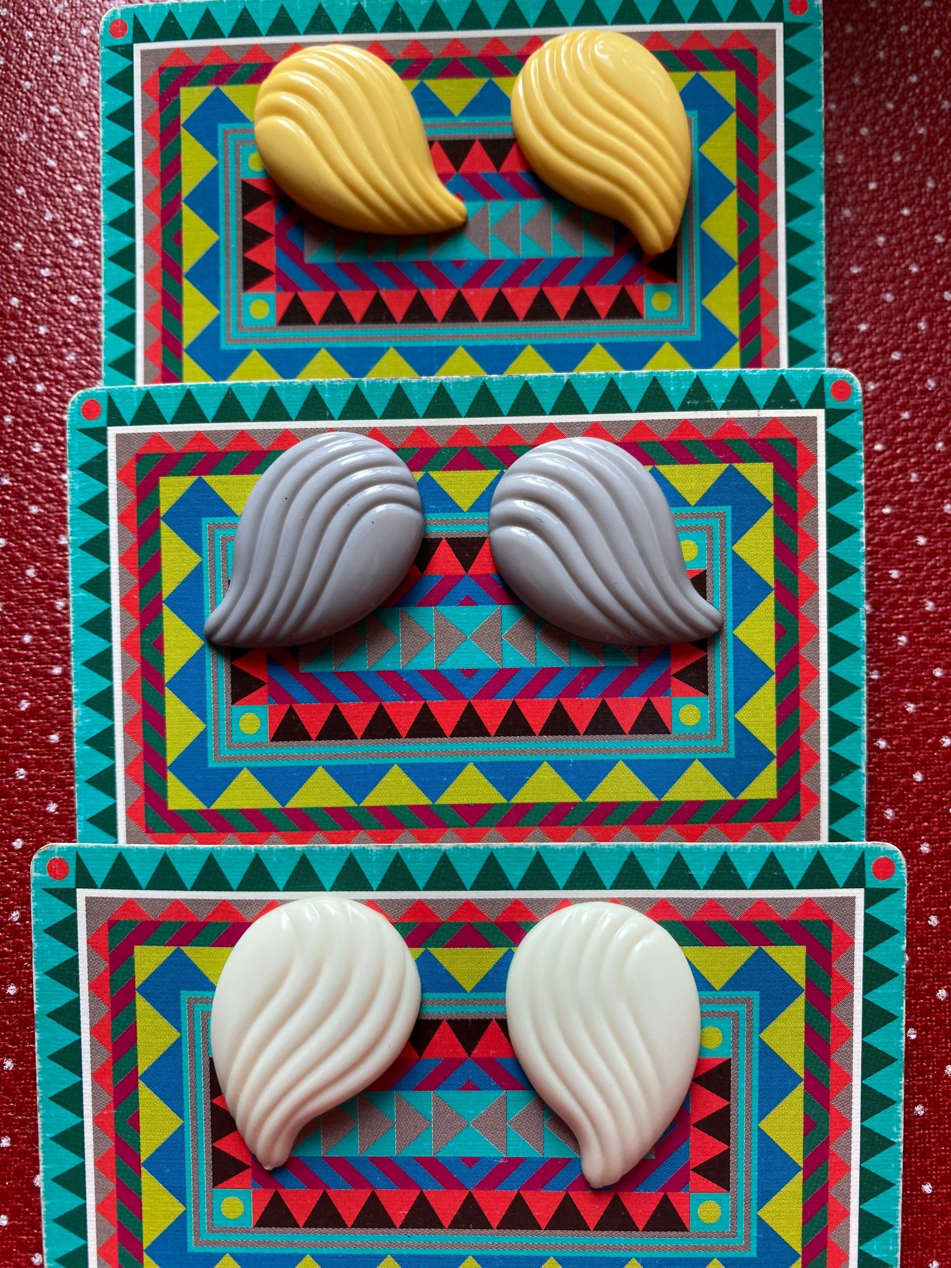 Three pairs of 80s earrings on colorful geometric-patterned cards against a red textured background.