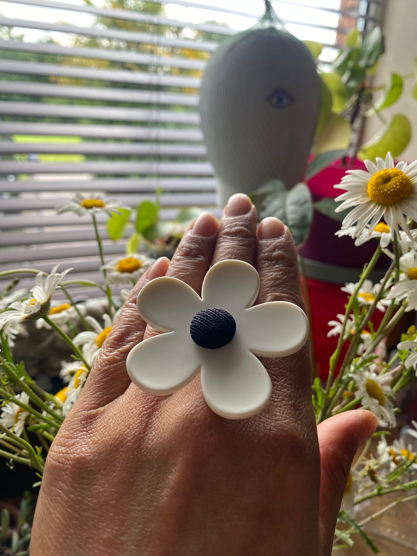 Hand holding a white flower-shaped object with a black center, surrounded by plants and a window.