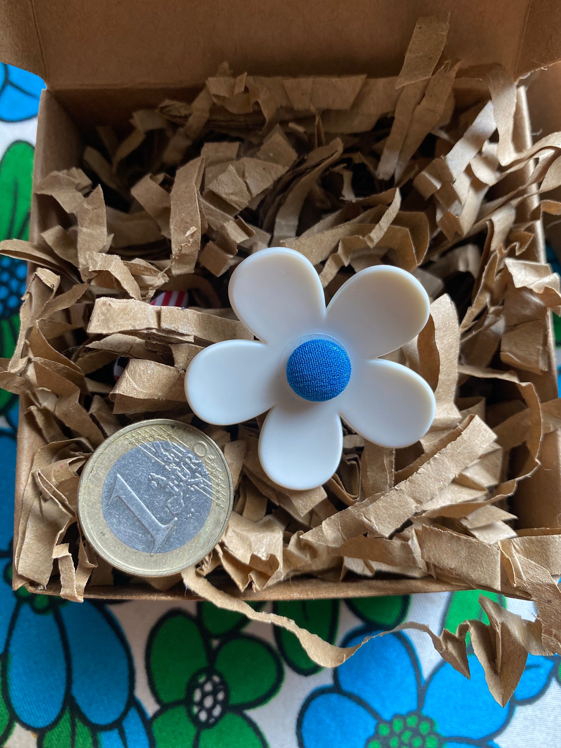 White handmade daisy flower-shaped ring with a blue center in a box of brown paper 
