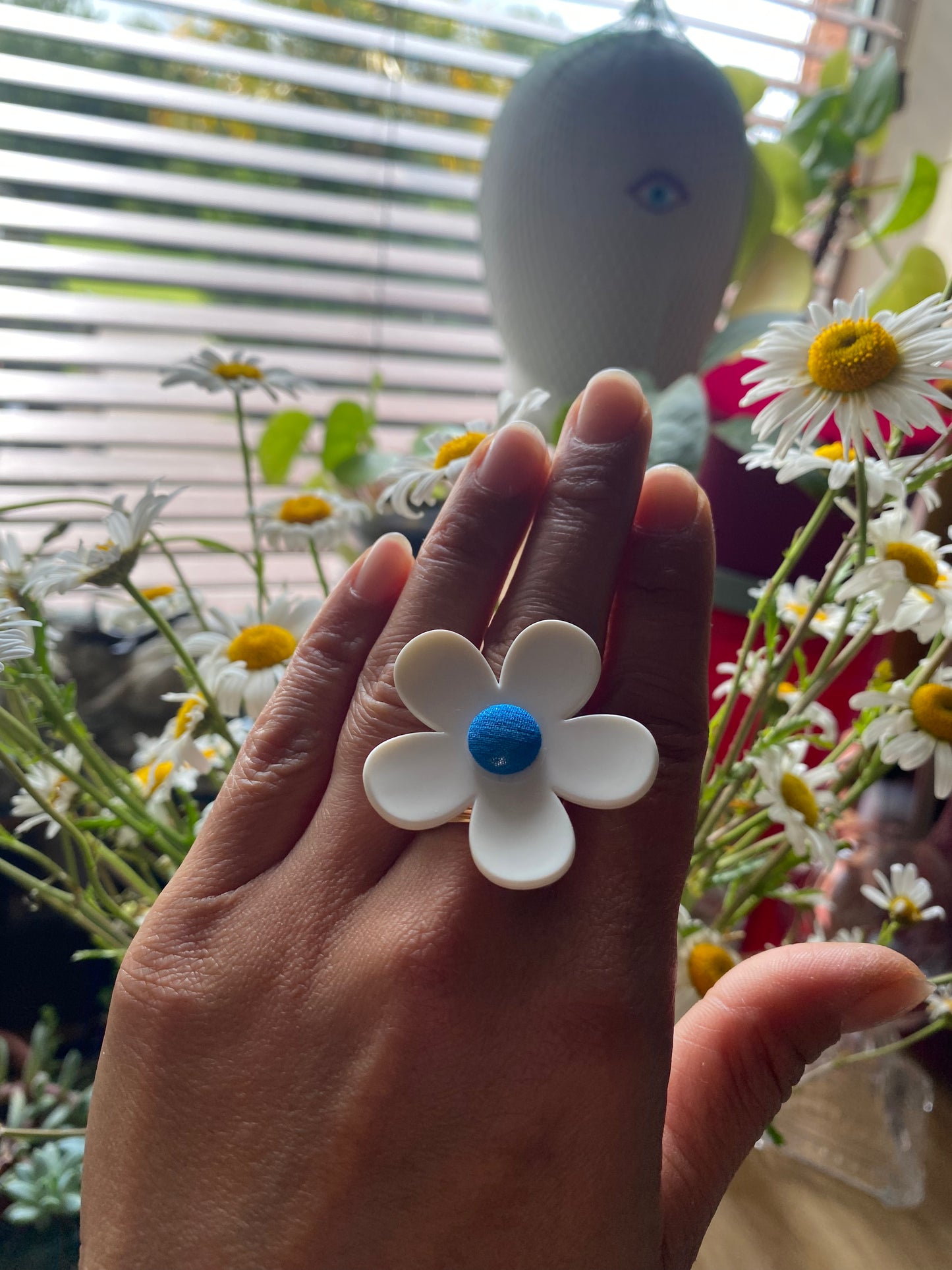 Hand wearing a white daisy flower-shaped ring with a blue center, surrounded by flowers.