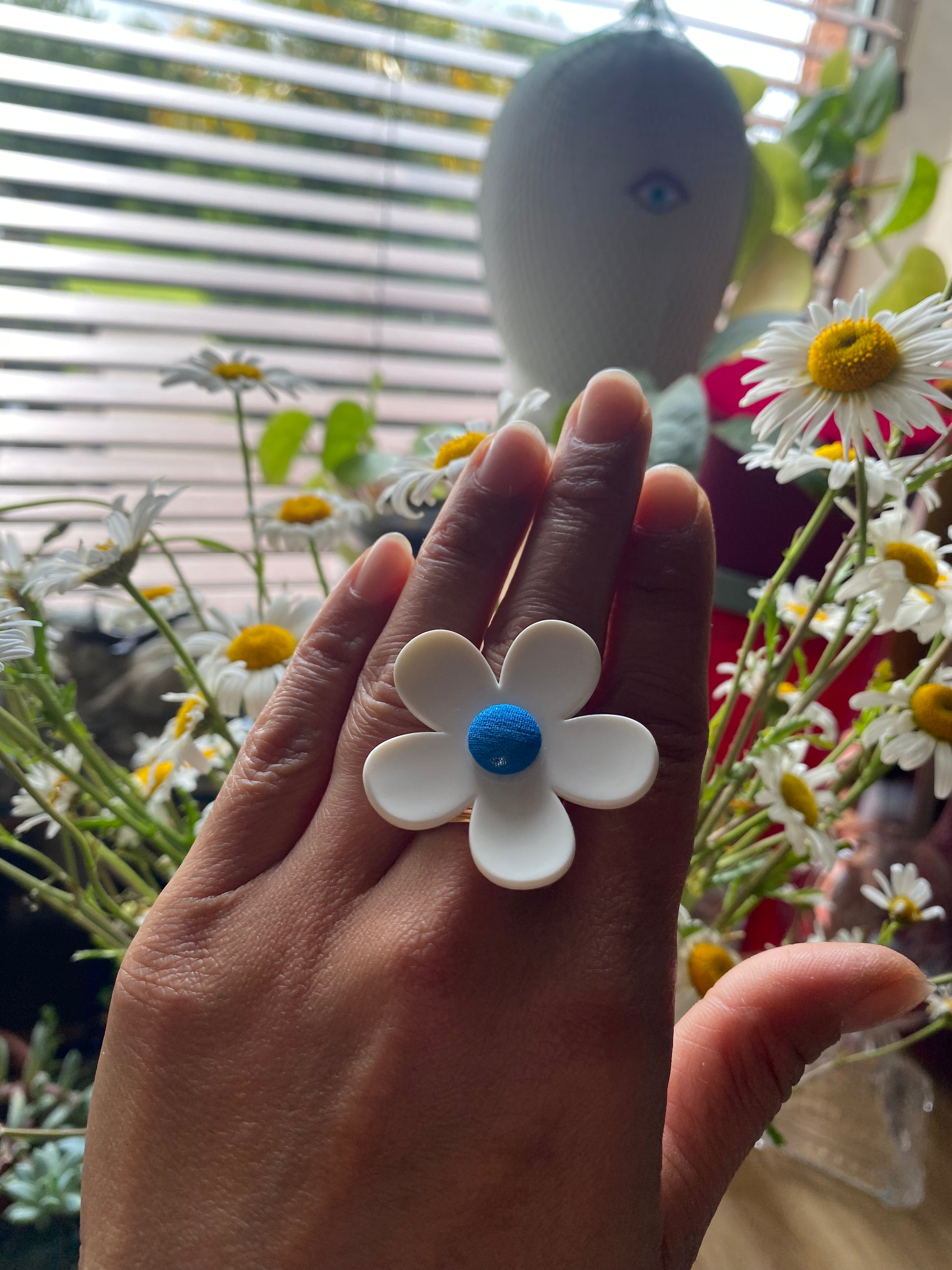 Hand wearing a white daisy flower-shaped ring with a blue center, surrounded by flowers.