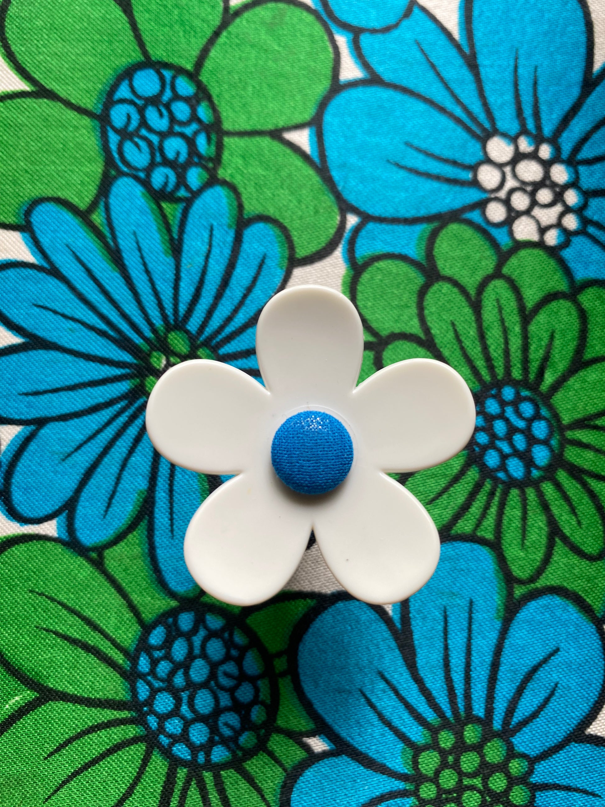 White daisy handmade flower with blue center on a fabric background with blue and green flowers