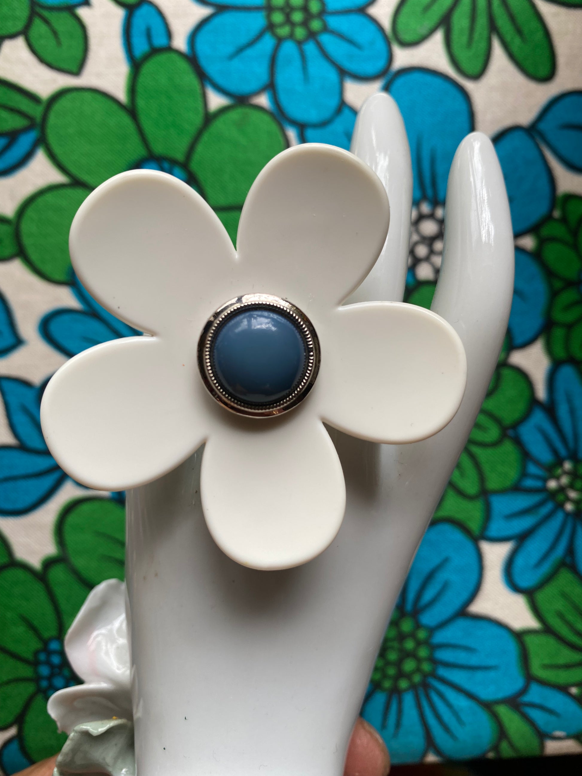 White ceramic hand modelling a handmade daisy flower-shaped ring with a blue center on a retro  floral-patterned background