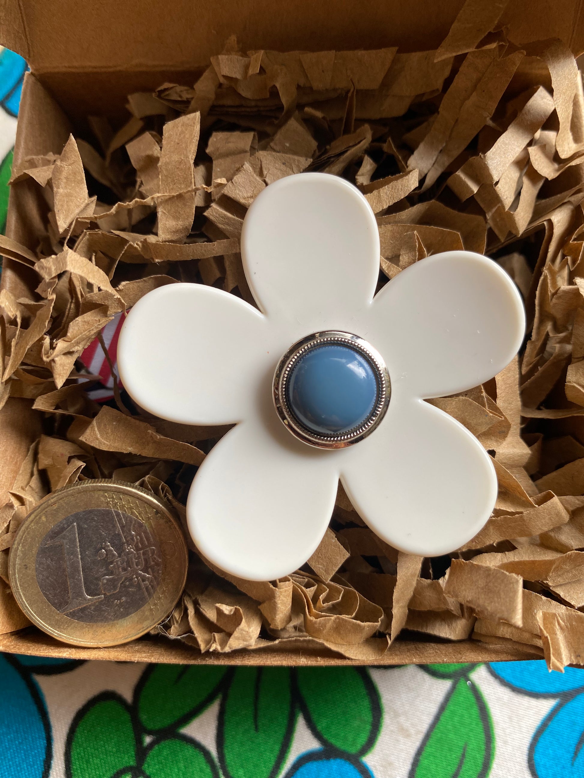 White flower-shaped ring with a blue center in a box of brown paper