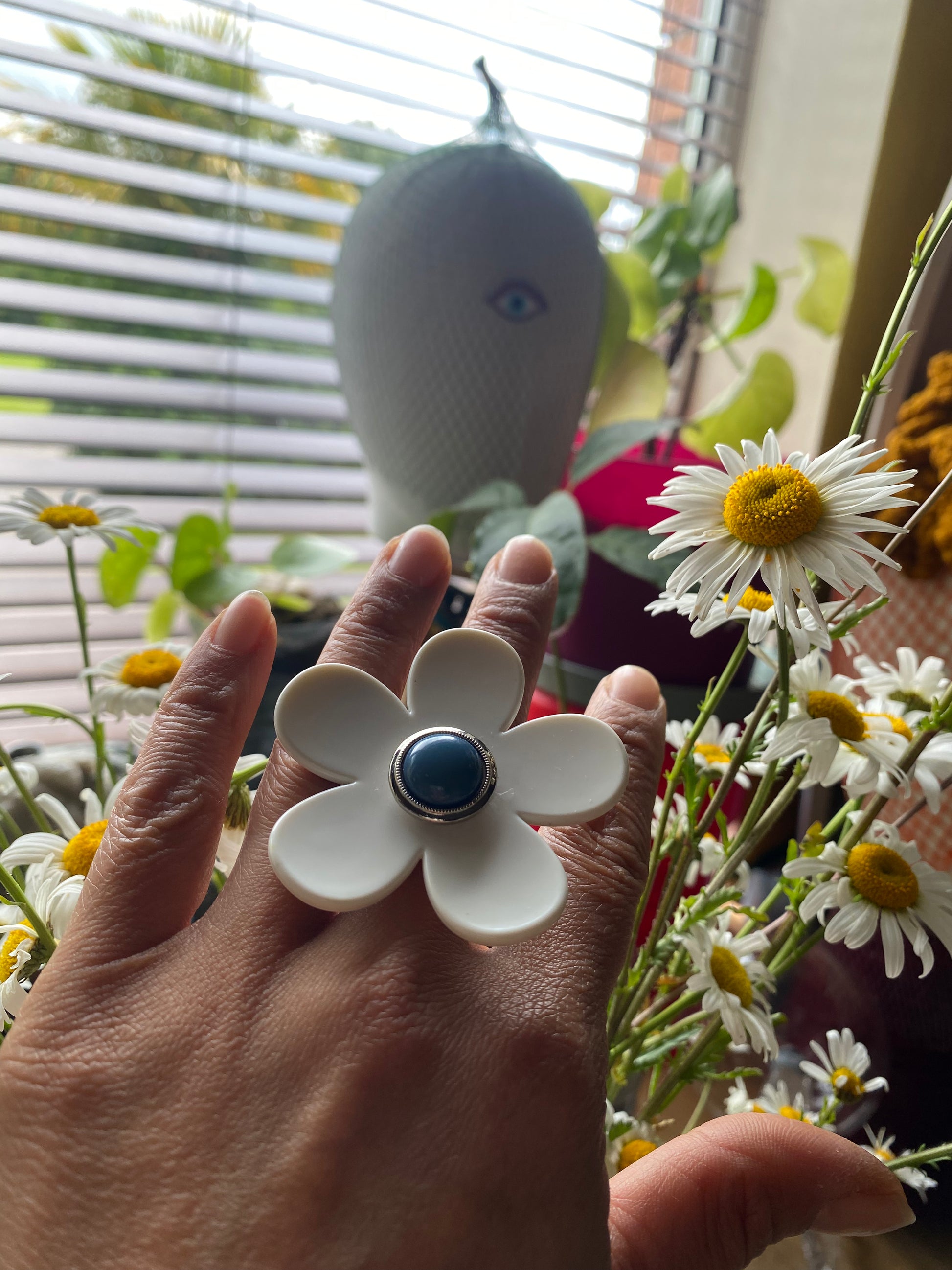Hand wearing a white flower-shaped ring with a blue center, surrounded by daisies.