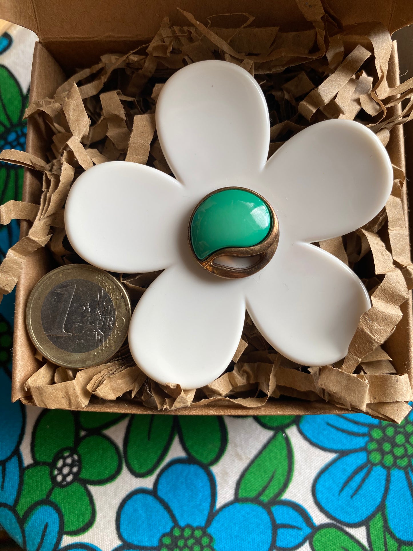 Handmade White daisy flower-shaped ring with a green center in a box with brown tissue paper, on a floral-patterned surface.