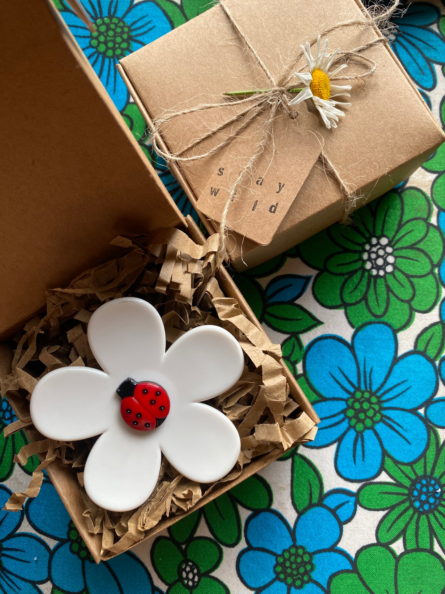 White up-cycled daisy flower-shaped ring with a red ladybug on a floral-patterned surface