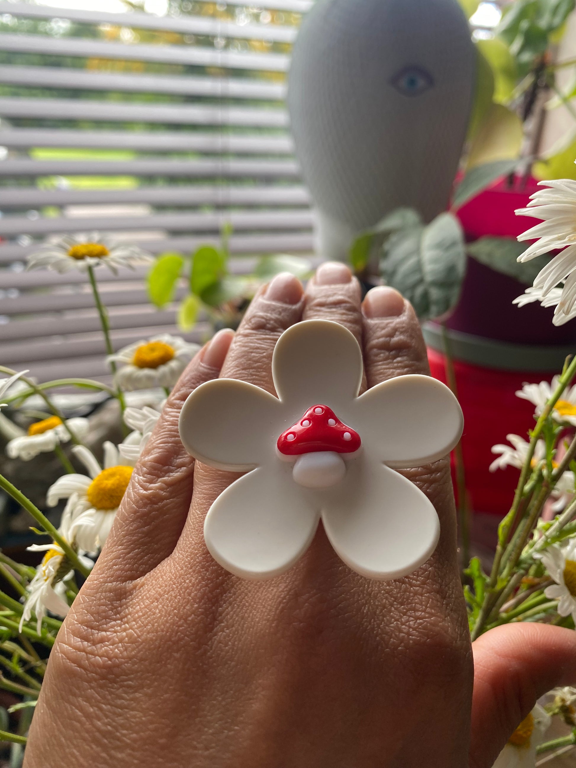 Hand wearing a handmade white daisy flower-shaped ring with a red mushroom center, surrounded by flowers and plants.
