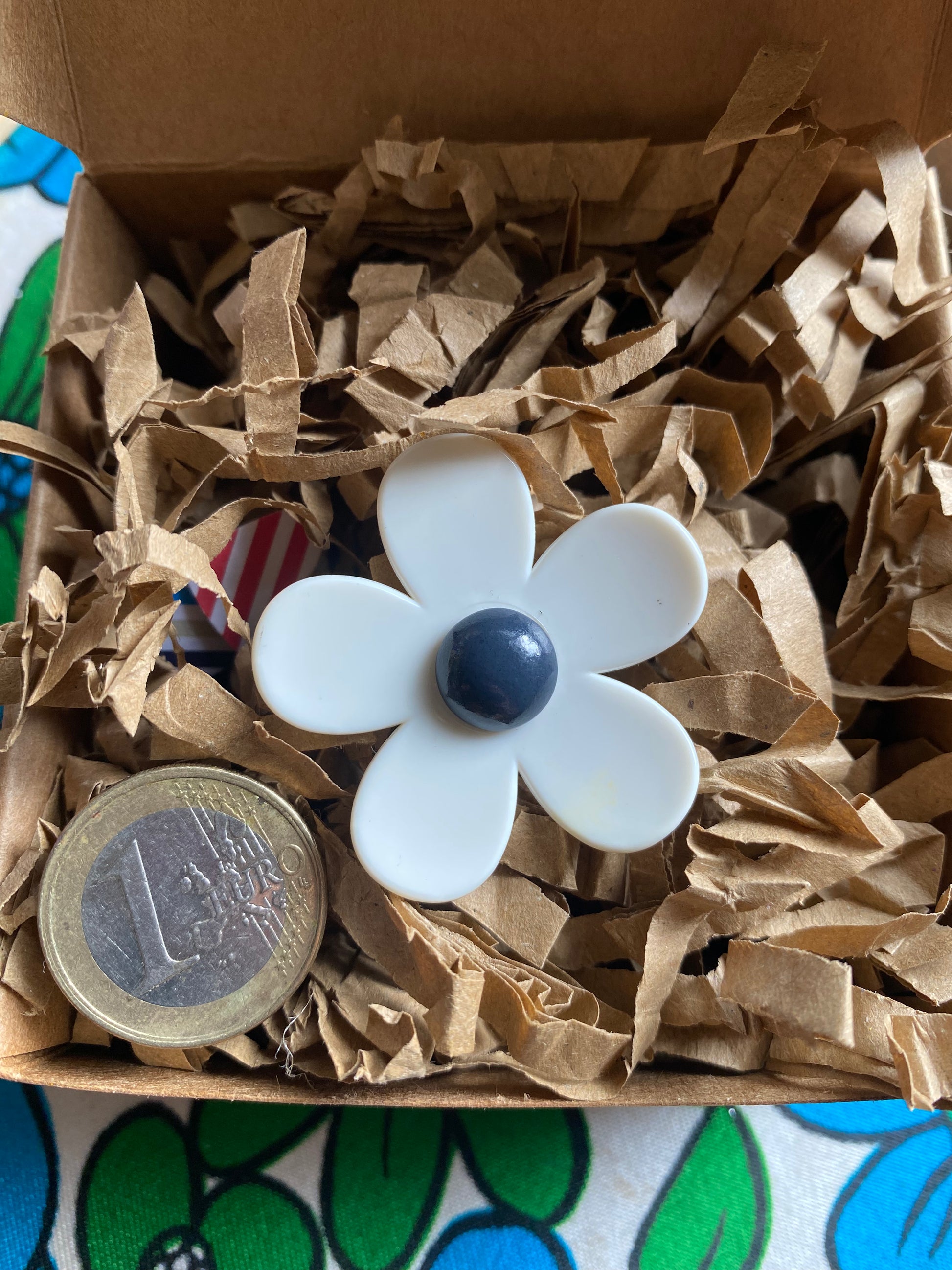 White handmade daisy flower-shaped ring with a blue center in a eco friendly box with brown tissue paper, next to a €1 coin for size.