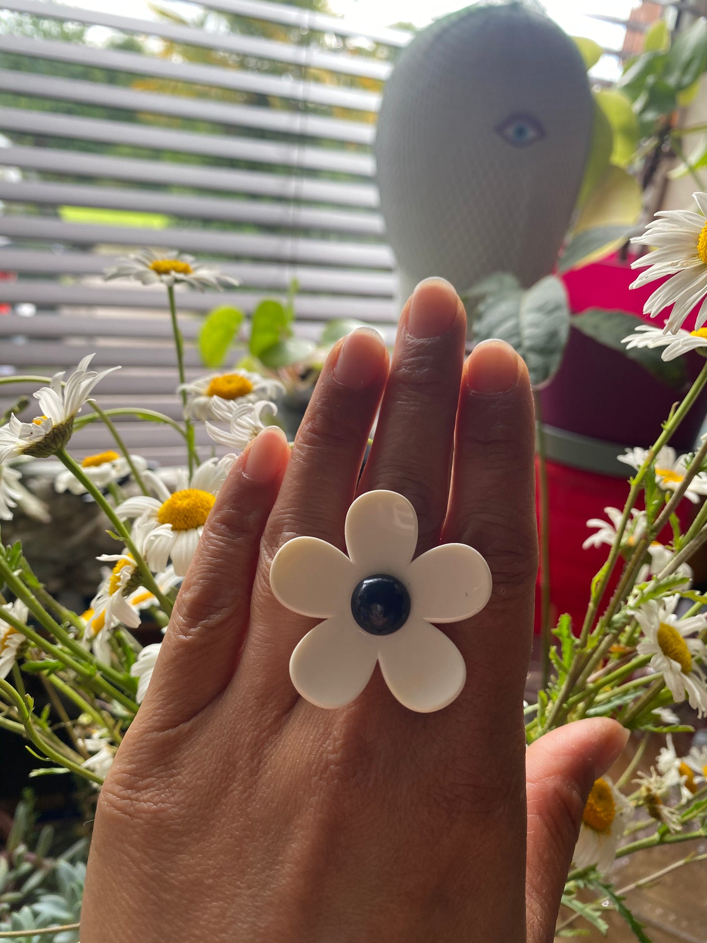 Hand wearing a white handmade daisy flower-shaped ring with a navy center, surrounded by flowers.