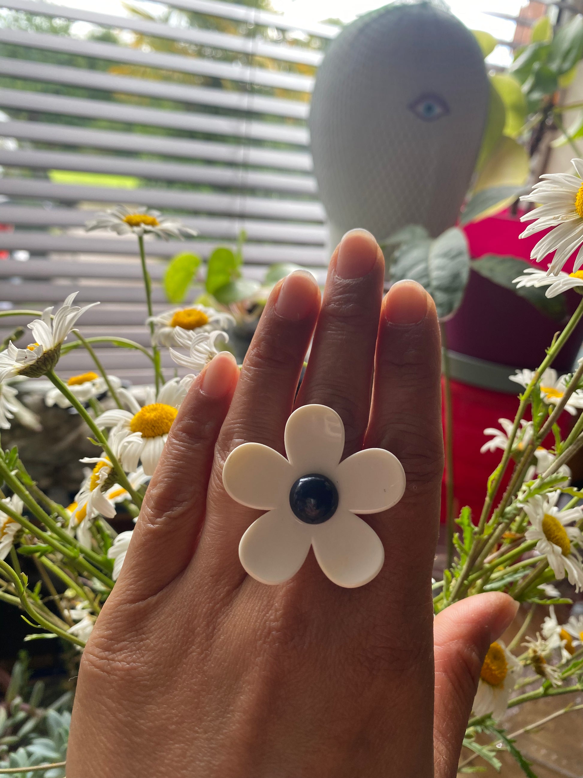 Hand wearing a white handmade daisy flower-shaped ring with a navy center, surrounded by flowers.