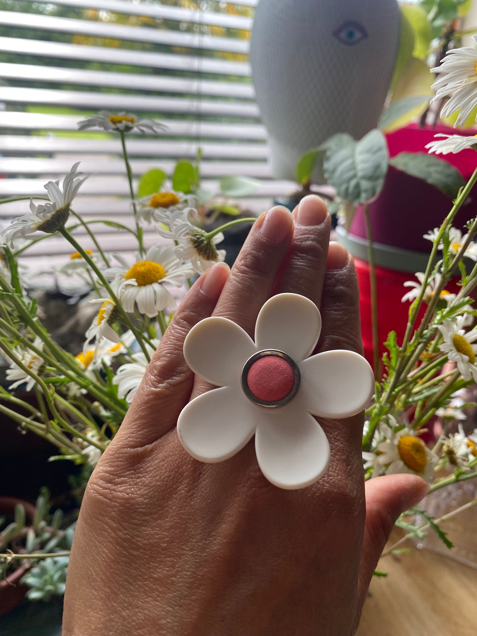 Hand holding a handmade white daisy flower-shaped ring with a pink center, surrounded by greenery and flowers.