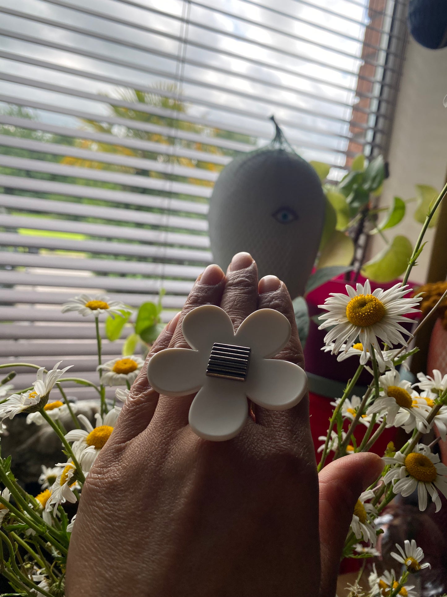 Hand wearing a handmade upcycled white flower-shaped ring with a blurred background of plants and a window.