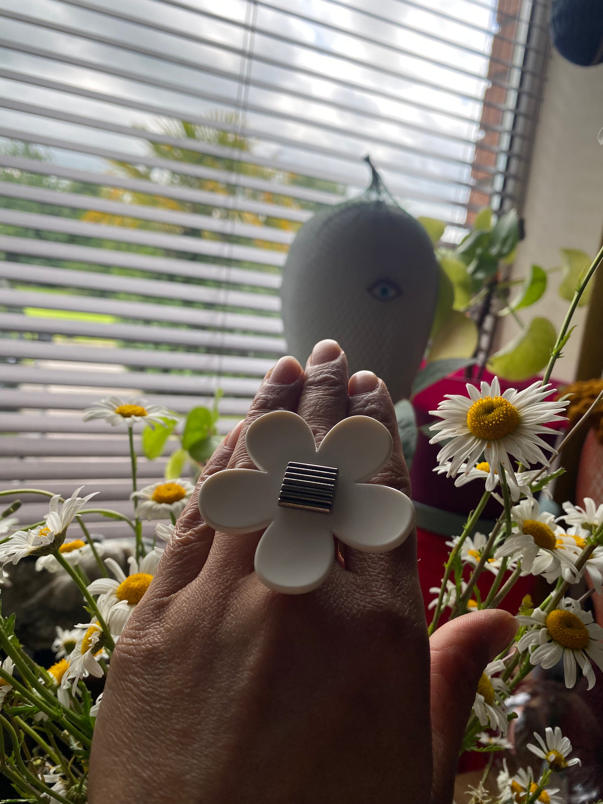 Hand wearing a handmade upcycled white flower-shaped ring with a blurred background of plants and a window.