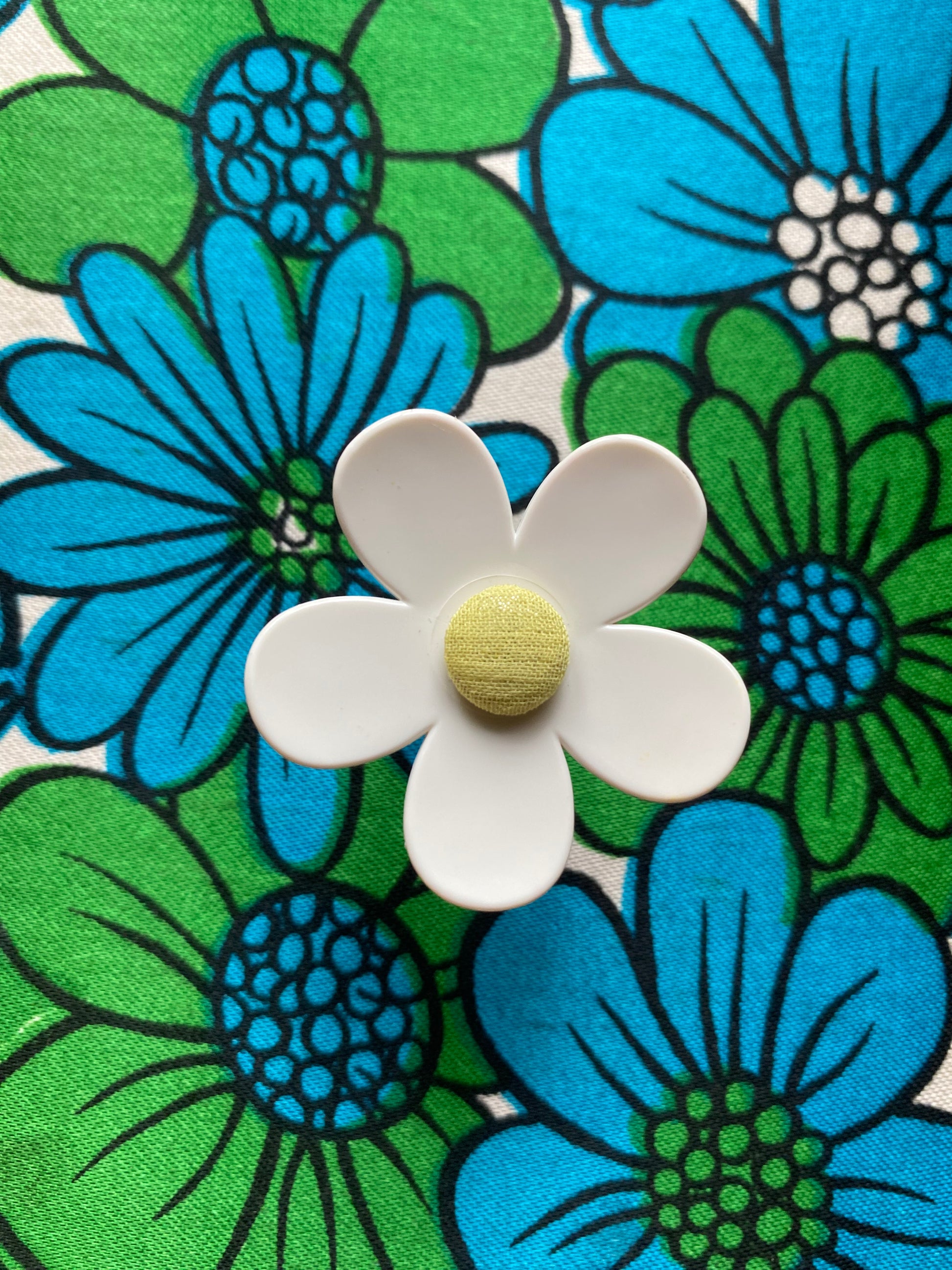 Handmade white daisy flower with a yellow center on a fabric background with blue and green floral retro patterns.