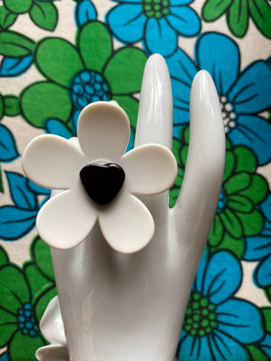 White ceramic hand modelling a handmade daisy ring with a black heart shaped in the middle on a floral retro background