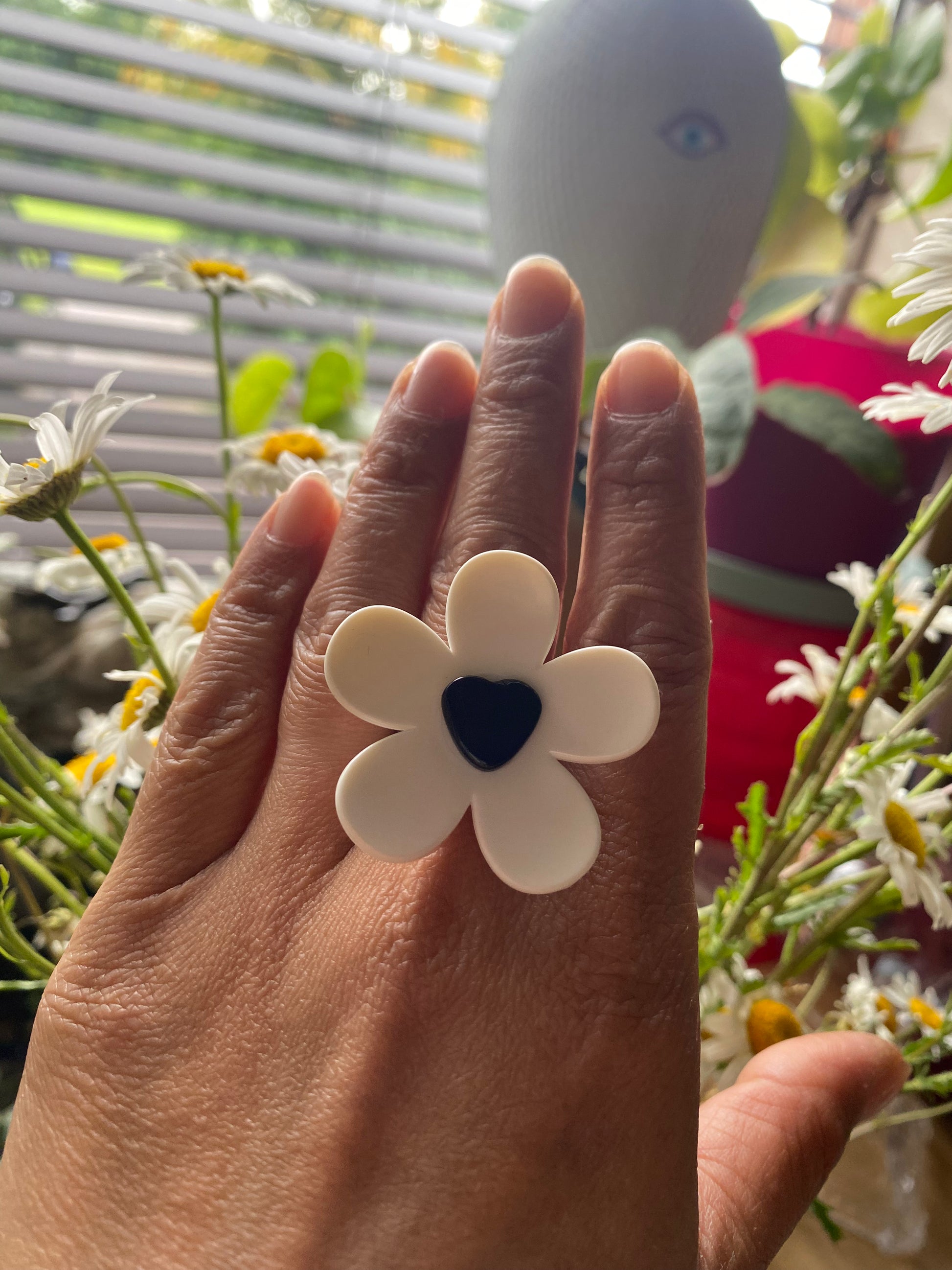 Hand wearing a white handmade daisy flower-shaped ring with a black center, surrounded by flowers and plants.
