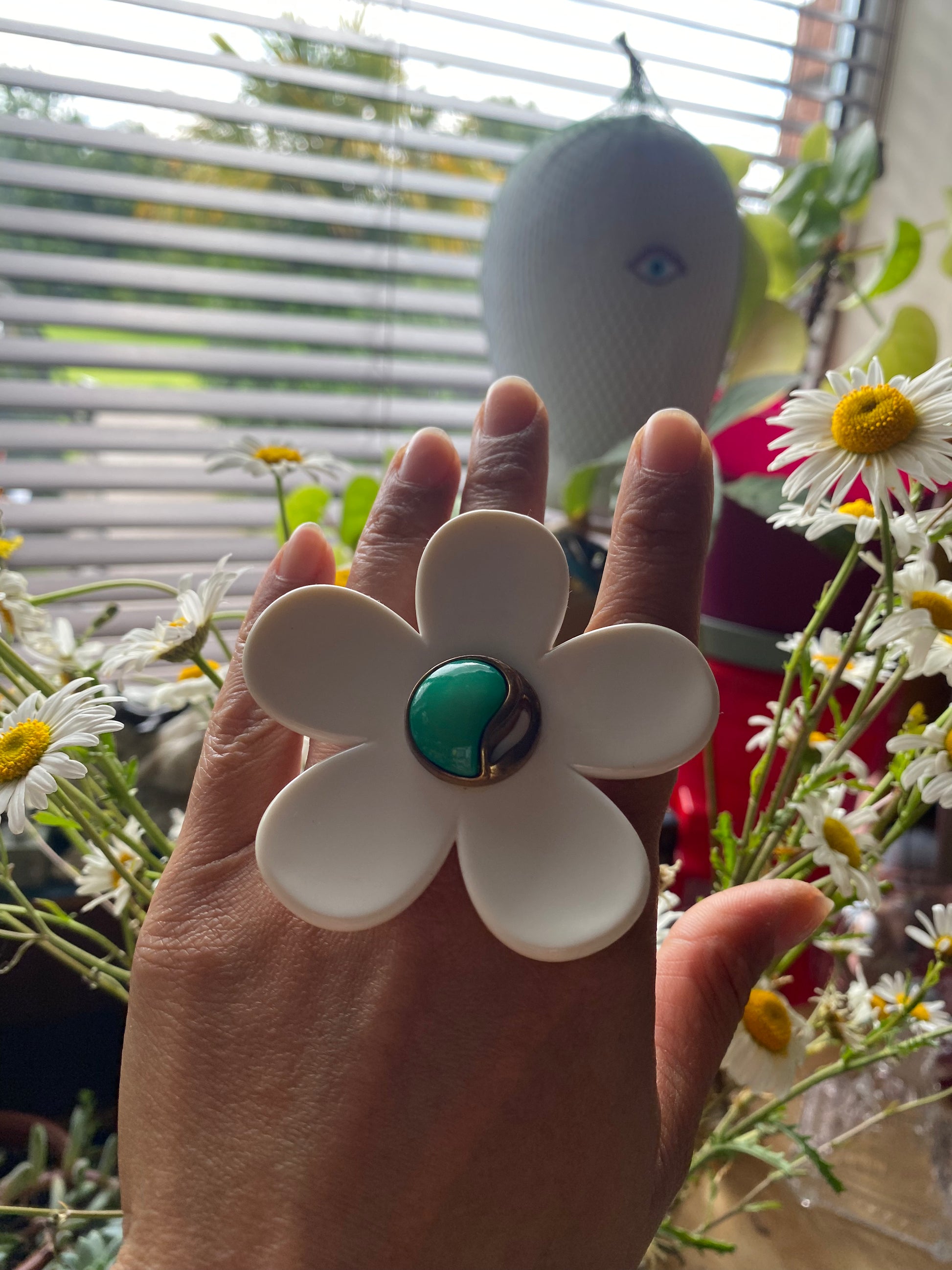 Hand modelling a white handmade daisy flower-shaped ring with a green center in front of a window with blinds.